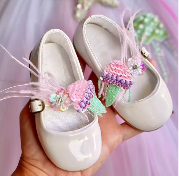 White children's shoes with decorative flowers held in a hand against a blurred background